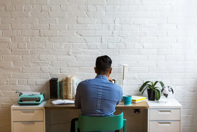 Back view of man at desk with typewriter and books, creating in a minimalist workspace. Essay research guide: questions, search paths, notes, and source judgment