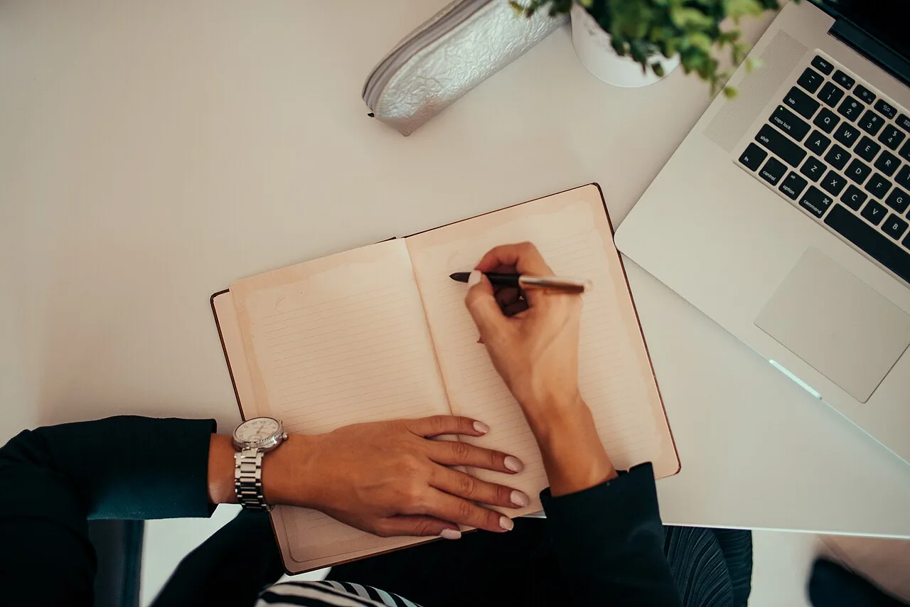 A person writing in a notebook beside a laptop